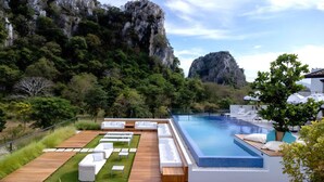 Zona d'una piscina exterior moderna, amb gandules blanques i un pont de fusta, envoltada de vegetació exuberant i penya-segades.