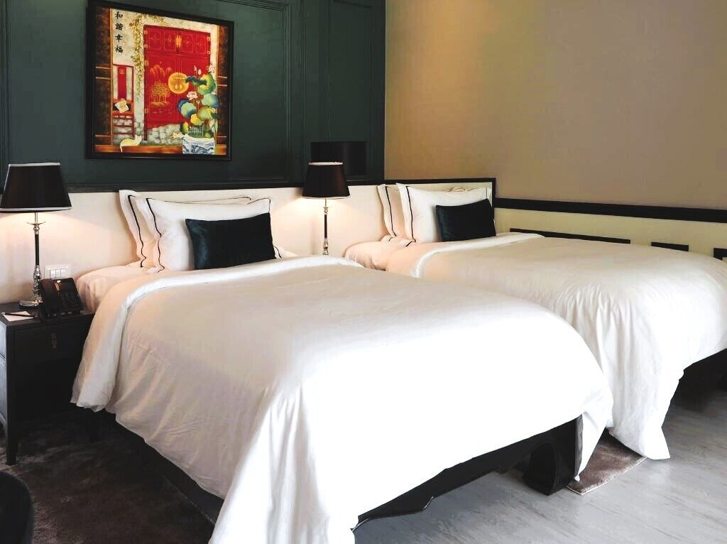 A hotel room with two beds, each with white bedding and black pillows, a nightstand with a lamp, and a painting on the wall.