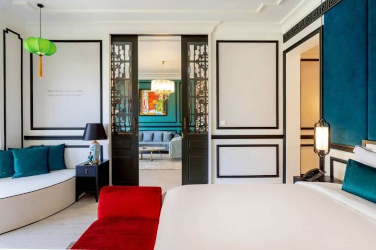 A modern hotel room with a large bed, a red accent chair, a seating area with a sofa and armchairs, and a view into the adjacent room.