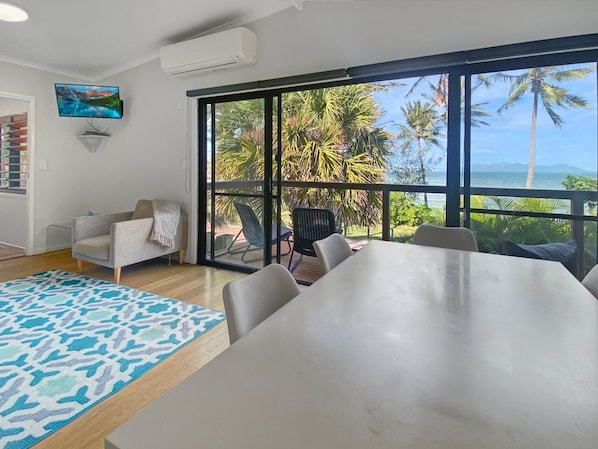 A modern room with a large table, a sofa, and a view of palm trees and the ocean.