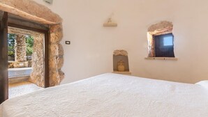 A bedroom with a stone wall, a bed with a patterned bedspread, a small shelf with a vase, and a window with a view of the outdoors.