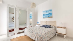 Une chambre à coucher avec un lit, une table de chevet, une commode et un tableau représentant la mer accroché au mur.
