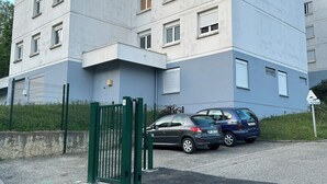 A multi-story building with a modern design, featuring a gated entrance and a parking area with cars.
