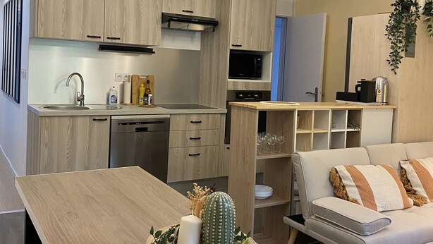 A modern kitchen with wooden cabinets, a built-in microwave, and a dining area with a light-colored table and sofa.