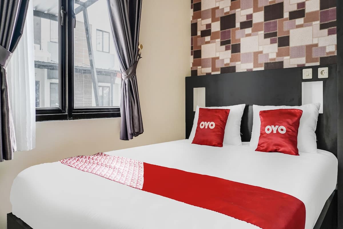 Une chambre d’hôtel avec un lit, deux oreillers rouges ornés du mot « OYO », une couvre-lit rouge et blanche à motifs, une fenêtre avec des rideaux et un mur décoré d’un motif géométrique.