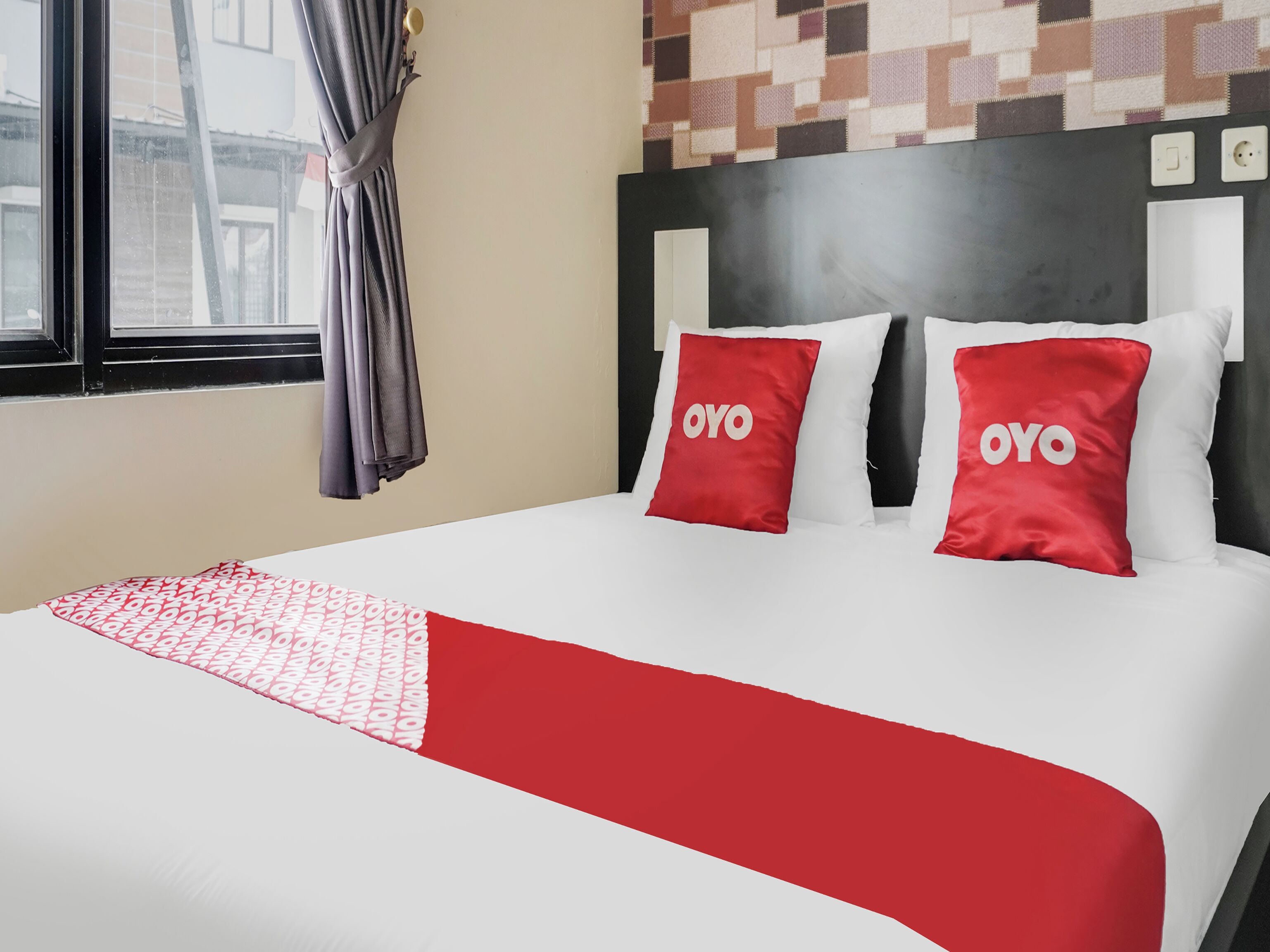A hotel room with a bed, two pillows with 'OYO' branding, a patterned throw, a window with curtains, and a wall with a geometric pattern.