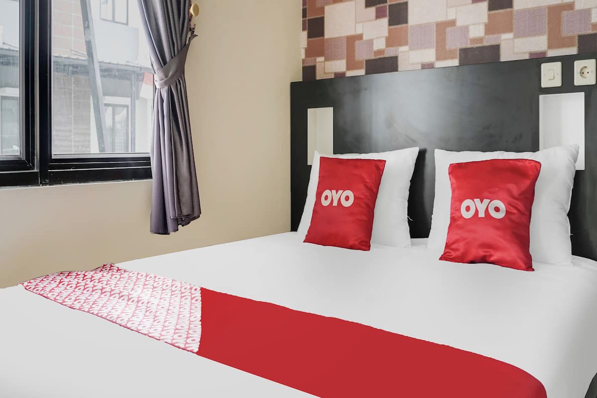 Une chambre d’hôtel avec un lit, deux oreillers portant la marque « OYO », un plaid à motifs, une fenêtre avec des rideaux et un mur orné d’un motif géométrique.