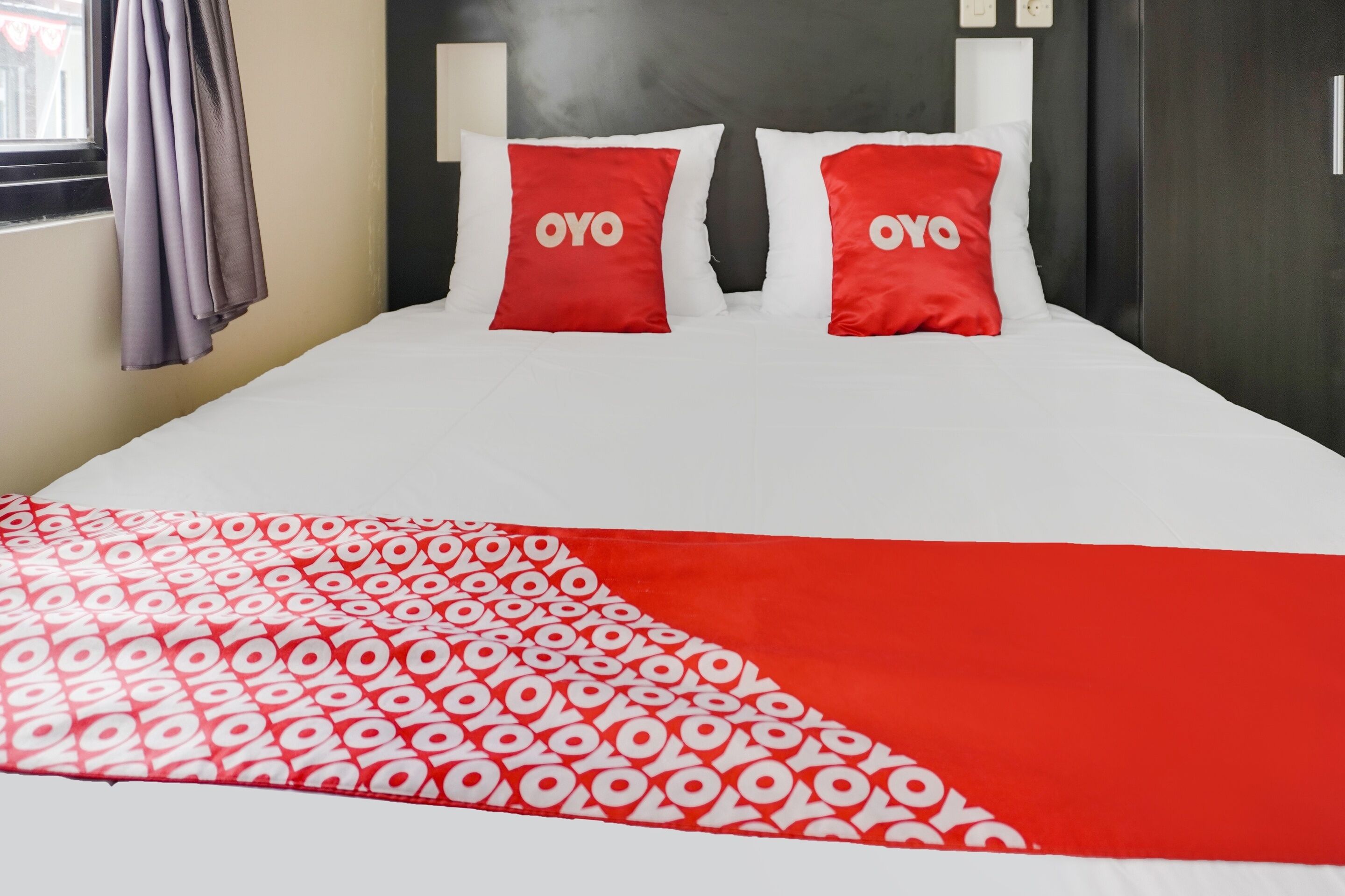 Ein Bett mit weißer und roter Bettwäsche, zwei rote Kissen mit dem Schriftzug „OYO“ sowie eine rote und weiße Decke mit Muster.