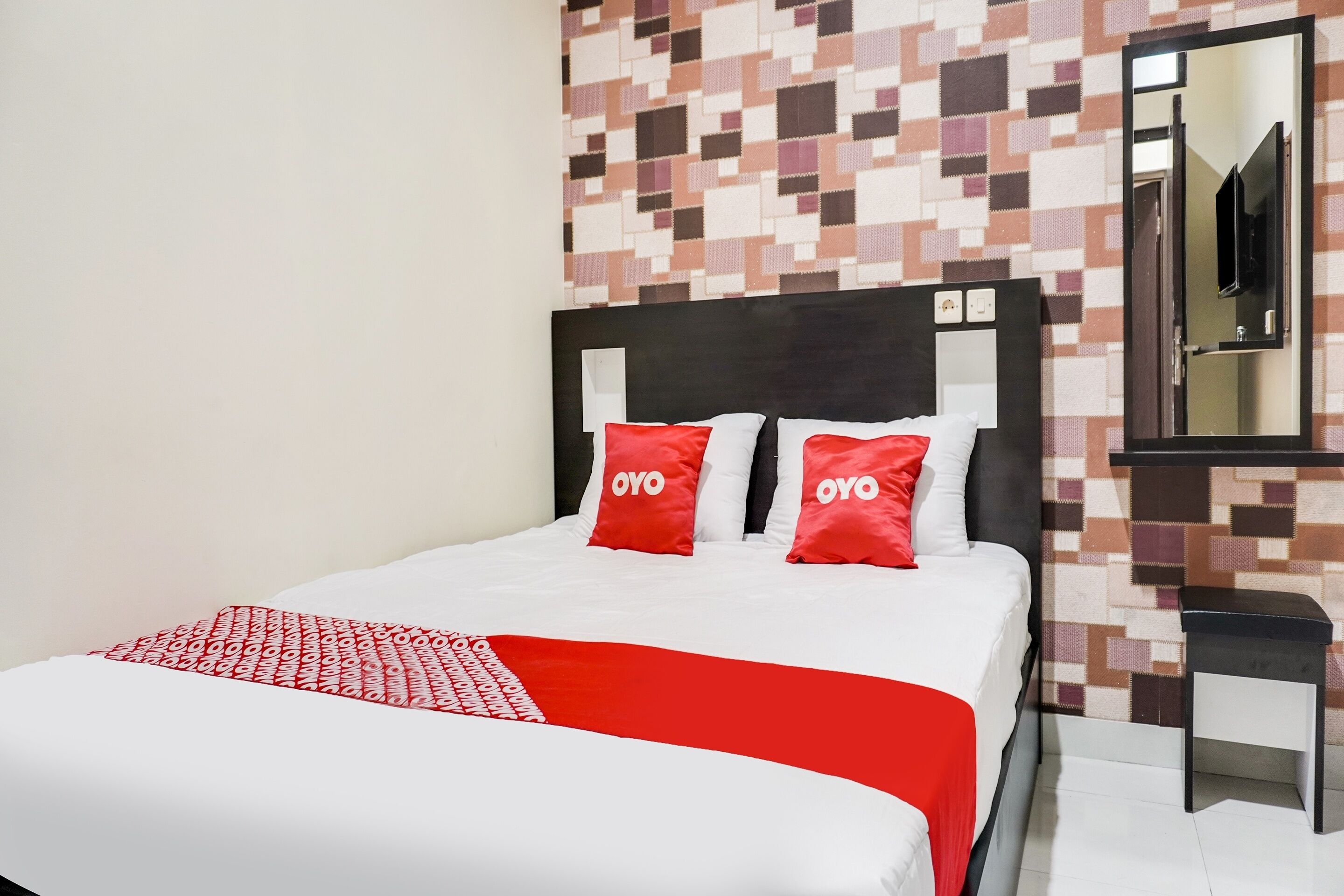 A hotel room with a bed, two red pillows with "OYO" text, a red and white blanket, a black nightstand, and a mirror on the wall.
