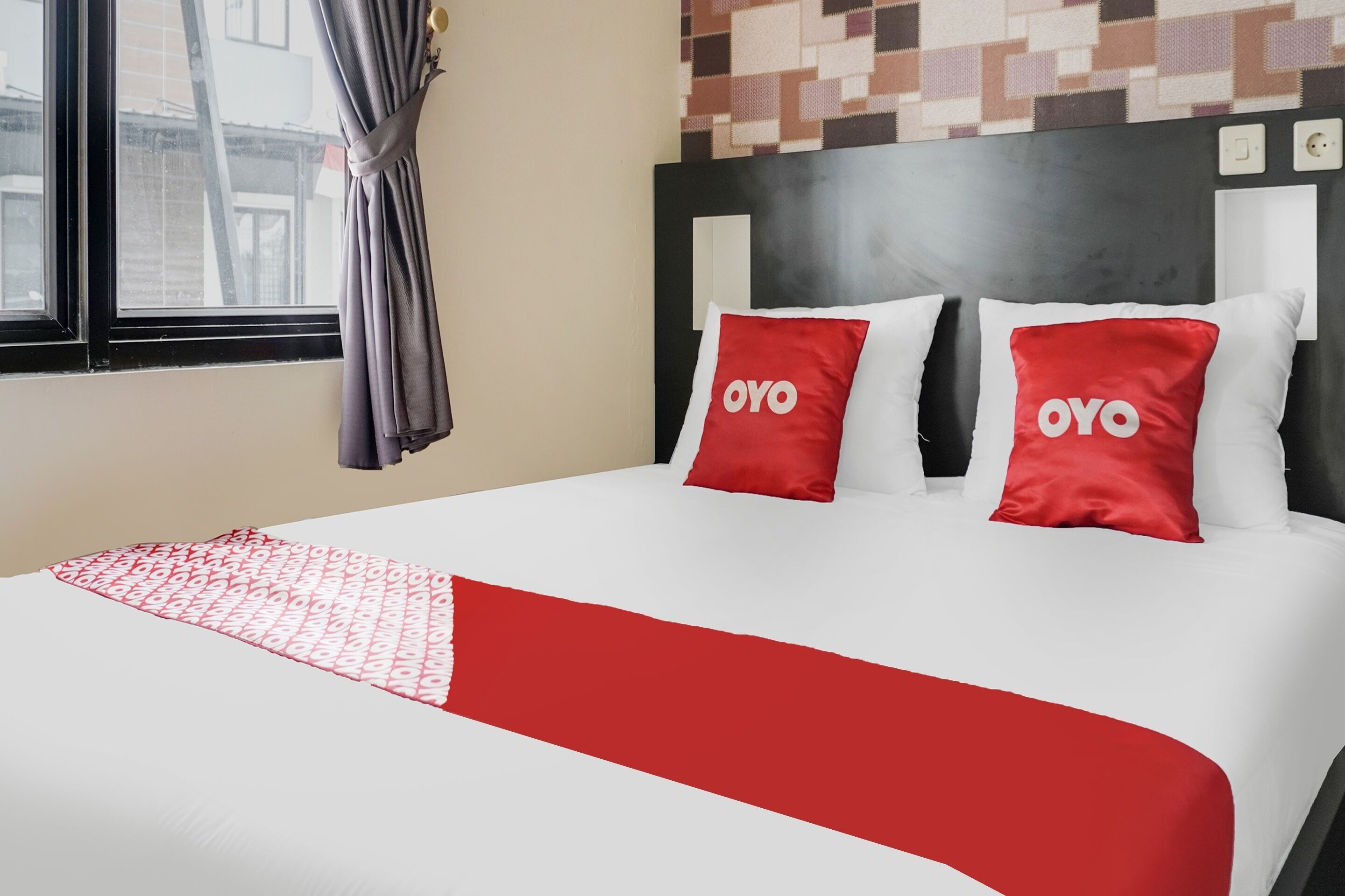 Habitación de hotel con cama, dos almohadas rojas con la marca 'OYO', una manta estampada y vistas a edificios a través de una ventana con cortinas.