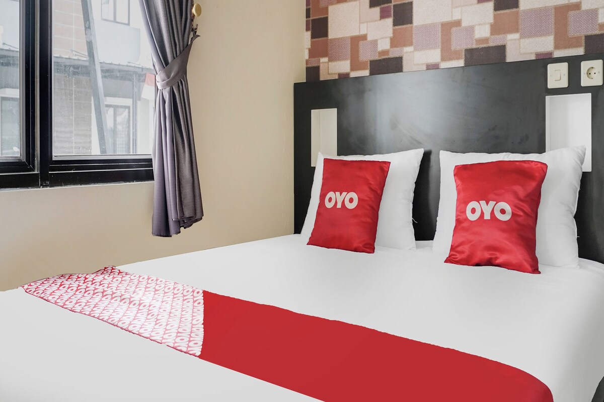 Une chambre d’hôtel avec un lit, deux oreillers portant la marque « OYO », un plaid à motifs, une fenêtre avec des rideaux et un mur orné d’un motif géométrique.