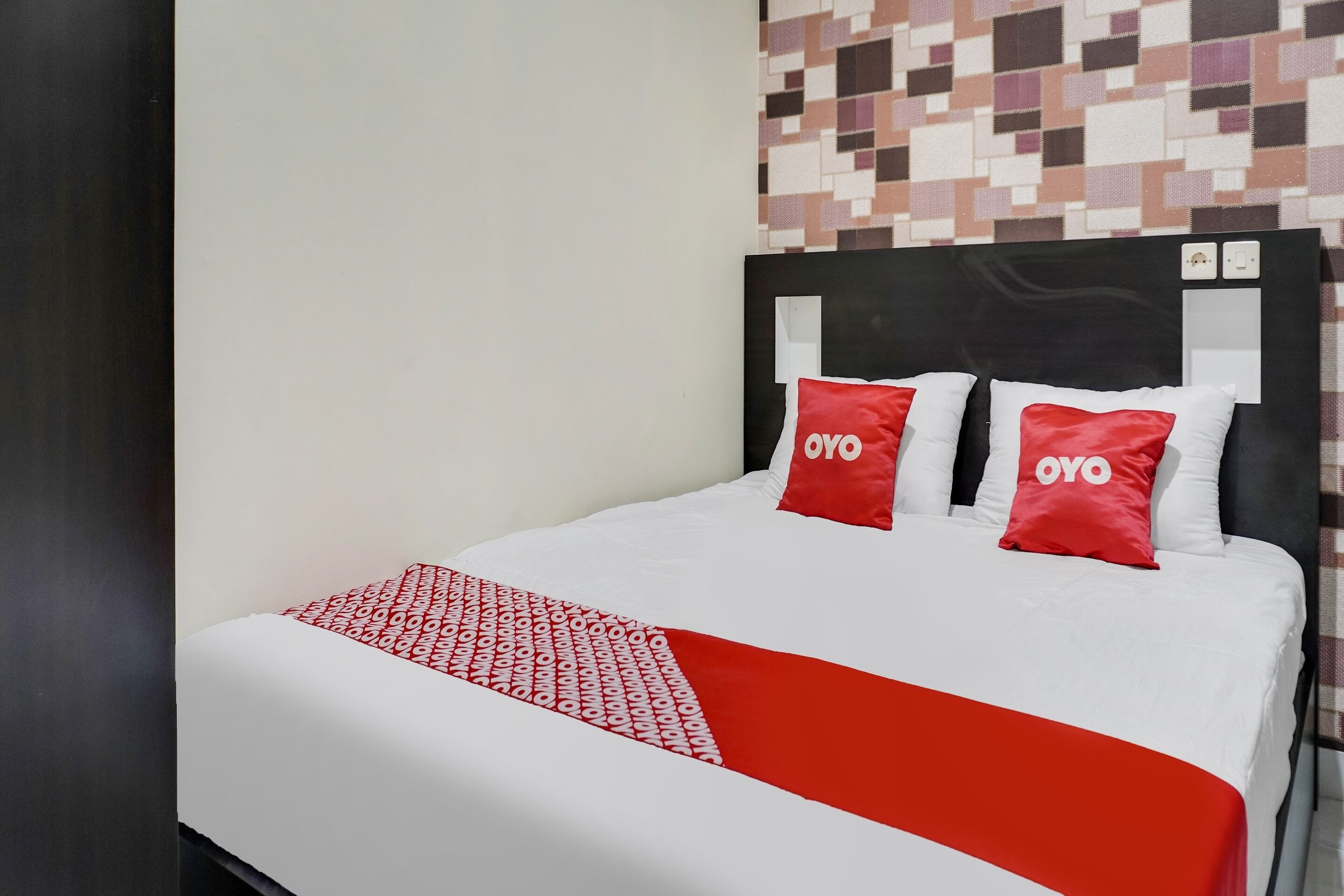 A bed with a white and red bedspread, two red pillows with "OYO" text, and a headboard with a black frame.
