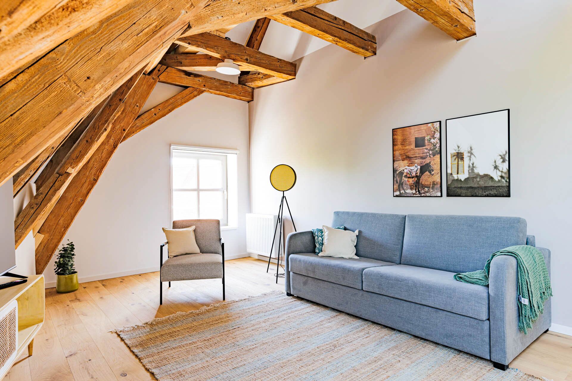 A cozy loft-style room with exposed wooden beams, a grey sofa, a small chair, and a floor lamp.