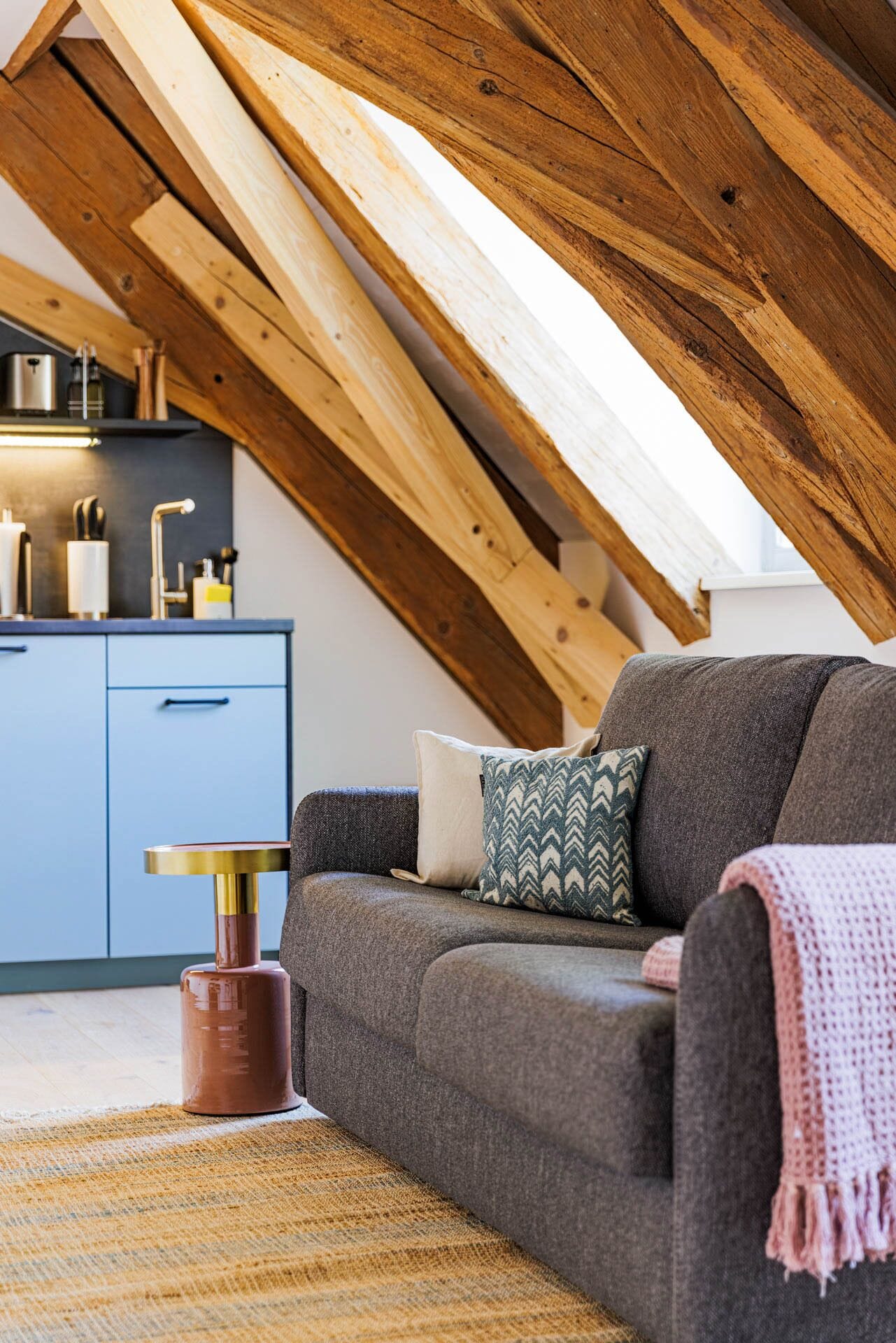 A loft-style room with a sloped ceiling, a grey sofa with patterned cushions, a small round table, and a woven rug.