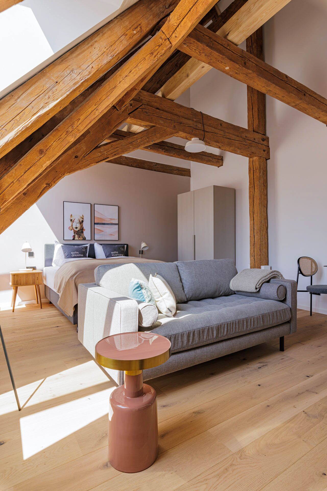 Dormitorio tipo loft con vigas de madera a la vista, un sofá gris, una mesa redonda y una cama con mesita de noche.