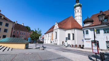 En brostensbelagt plads med historiske bygninger, en kirke med et kirketårn og en klar blå himmel.