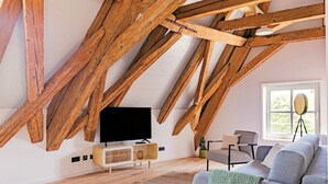 Une pièce de style « loft », avec des poutres en bois apparentes, un téléviseur posé sur un meuble en bois, un canapé gris et une fenêtre donnant sur un paysage verdoyant.