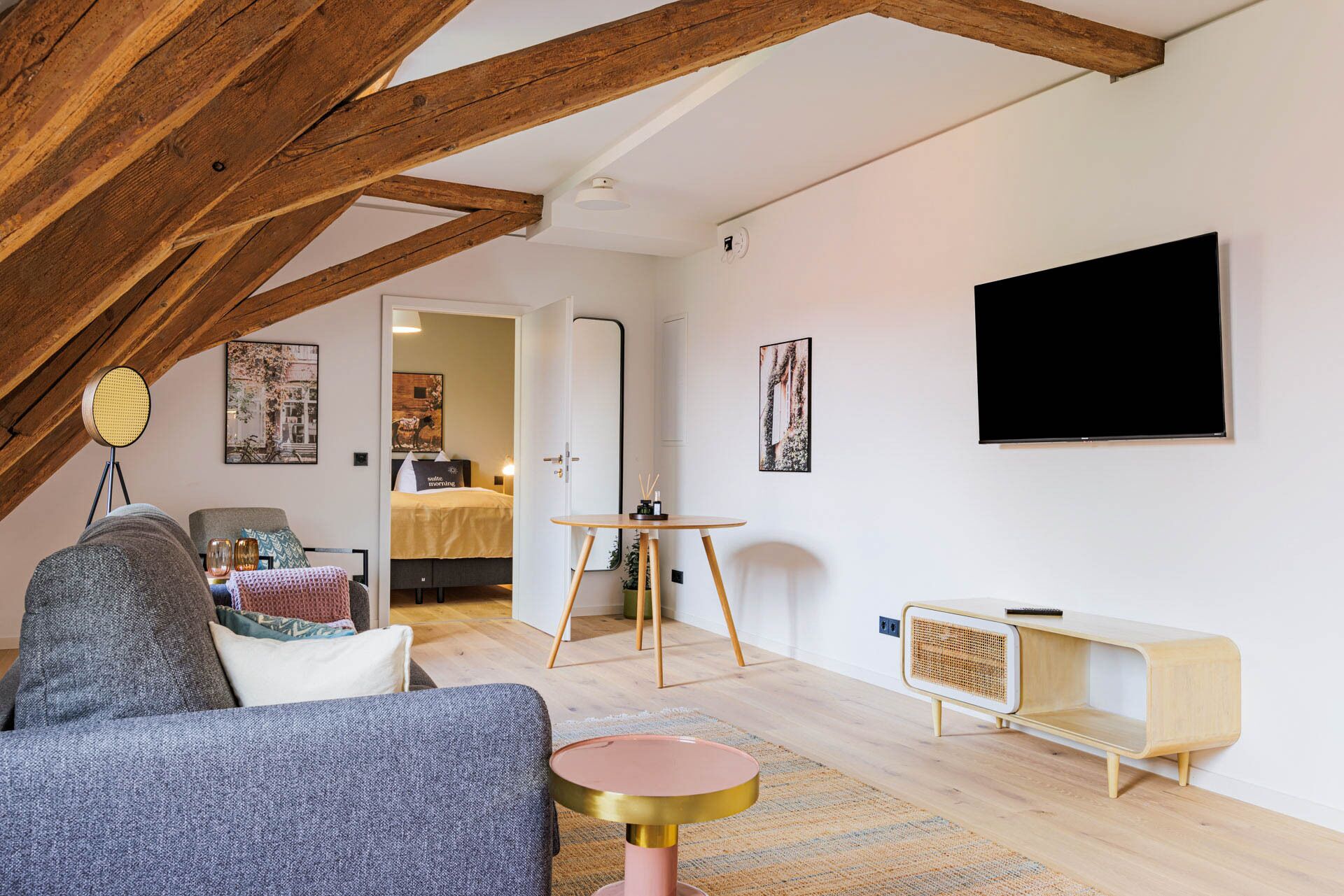 Ein gemütliches Schlafzimmer im Loft-Stil mit freiliegenden Holzbalken, einem Bett mit Kopfteil, einem kleinen runden Tisch, einem an der Wand befestigten Fernseher und einem Fenster mit Blick auf den Raum.