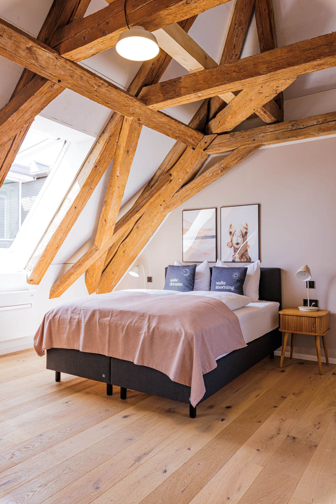 Un dormitorio con techo inclinado, vigas de madera, una cama con manta rosa, dos almohadas con texto, una mesita de noche con lámpara y dos cuadros en la pared.
