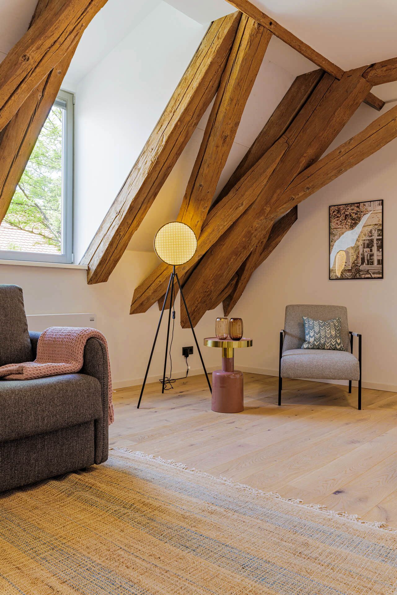 Ein Loft-Zimmer mit freiliegenden Holzbalken, einer modernen Stehlampe, einer Couch, einem Sessel und einem Teppich.