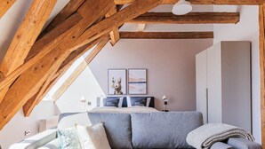 Une chambre de style « loft », avec des poutres en bois apparentes, un canapé gris et un lit doté d’une tête de lit blanche.