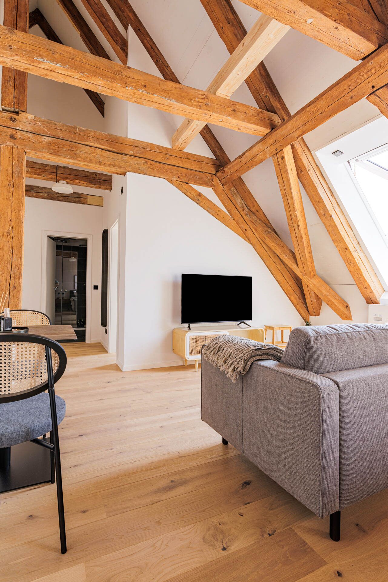 Ruime loft met zichtbare houten balken, een flatscreen televisie, een grijze bank en een eetgedeelte met een rieten stoel.
