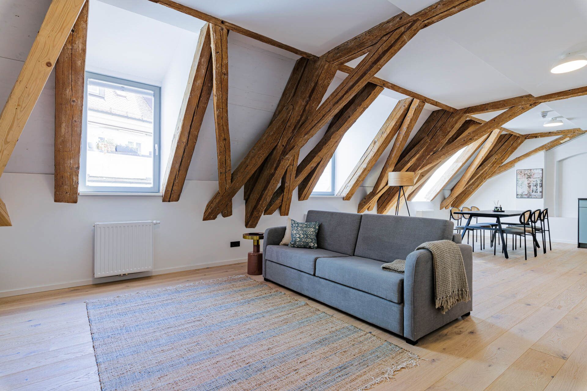 Een loft-stijl kamer met zichtbare houten balken, een grijze bank, een vloerkleed met patroon, en een eethoek met stoelen en een tafel.
