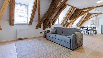Een loft-stijl kamer met zichtbare houten balken, een grijze bank, een vloerkleed met patroon, en een eethoek met stoelen en een tafel.