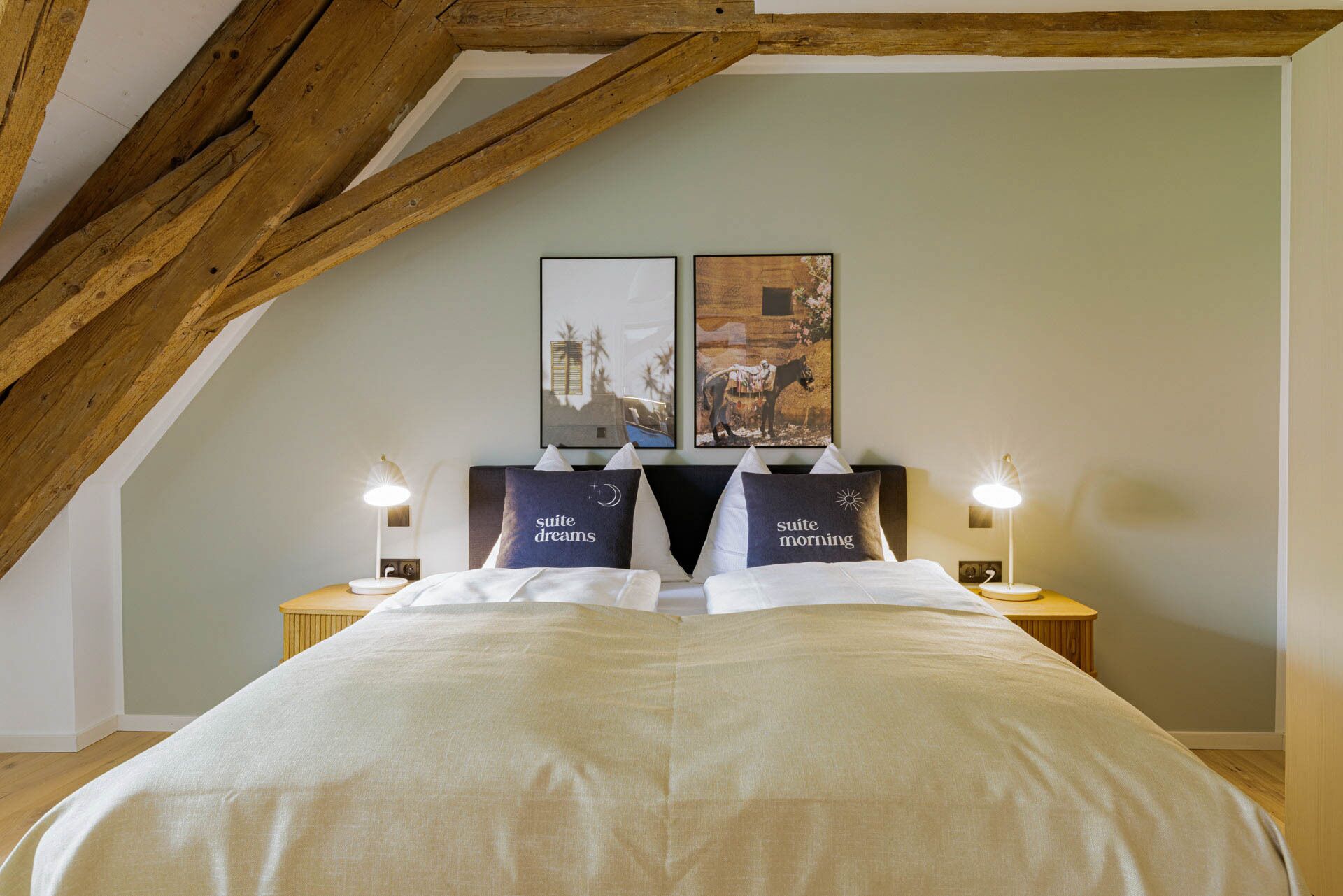 Un dormitorio con techo alto, vigas de madera, una cama grande con ropa blanca, dos mesitas de noche con lámparas y dos cuadros en la pared.