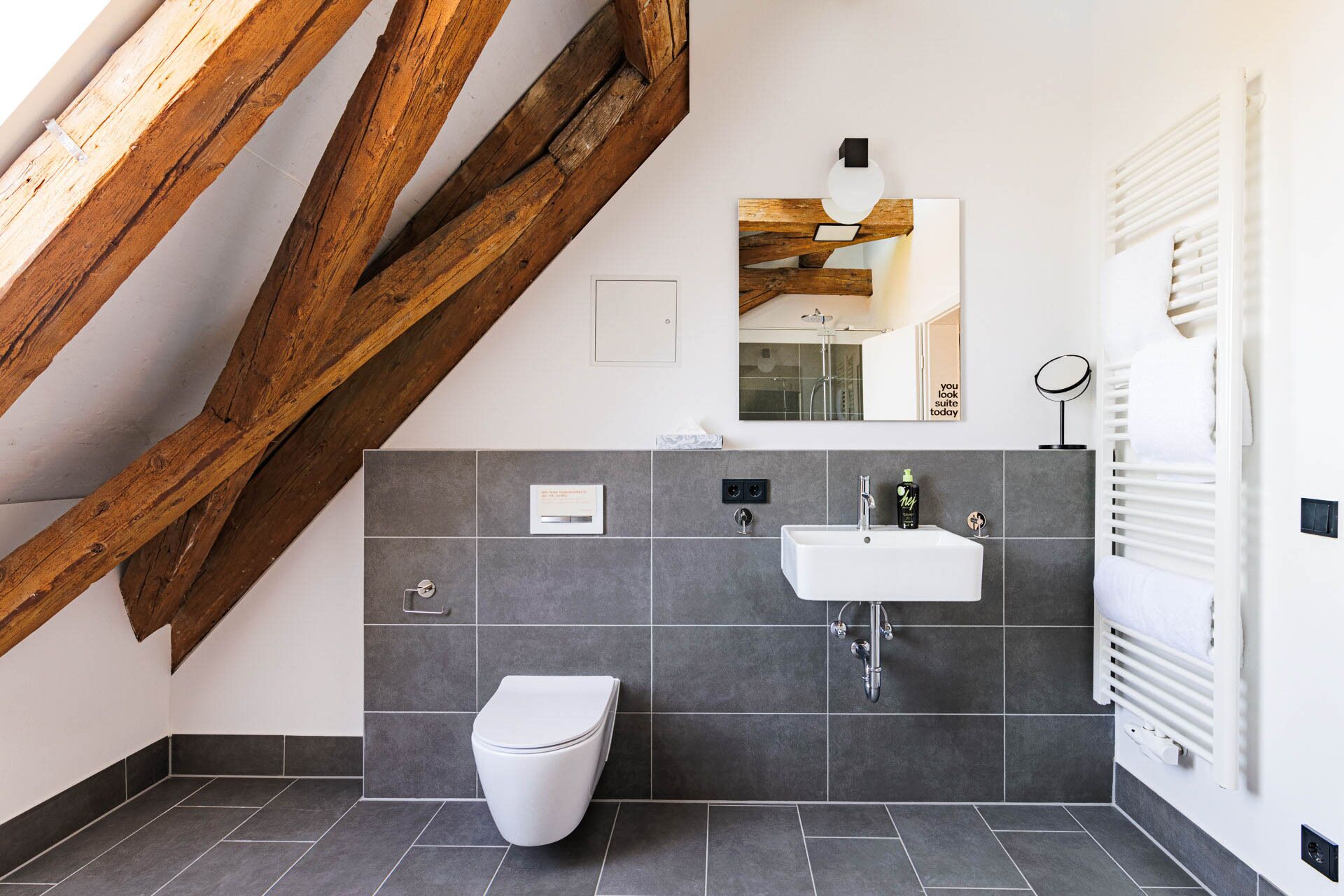 Un baño moderno con inodoro blanco, lavamanos y espejo. Se observa una estructura de vigas de madera sobre el inodoro, y un perchero a la derecha.