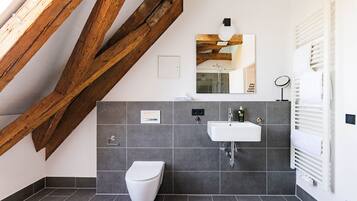 Un baño moderno con inodoro blanco, lavamanos y espejo. Se observa una estructura de vigas de madera sobre el inodoro, y un perchero a la derecha.