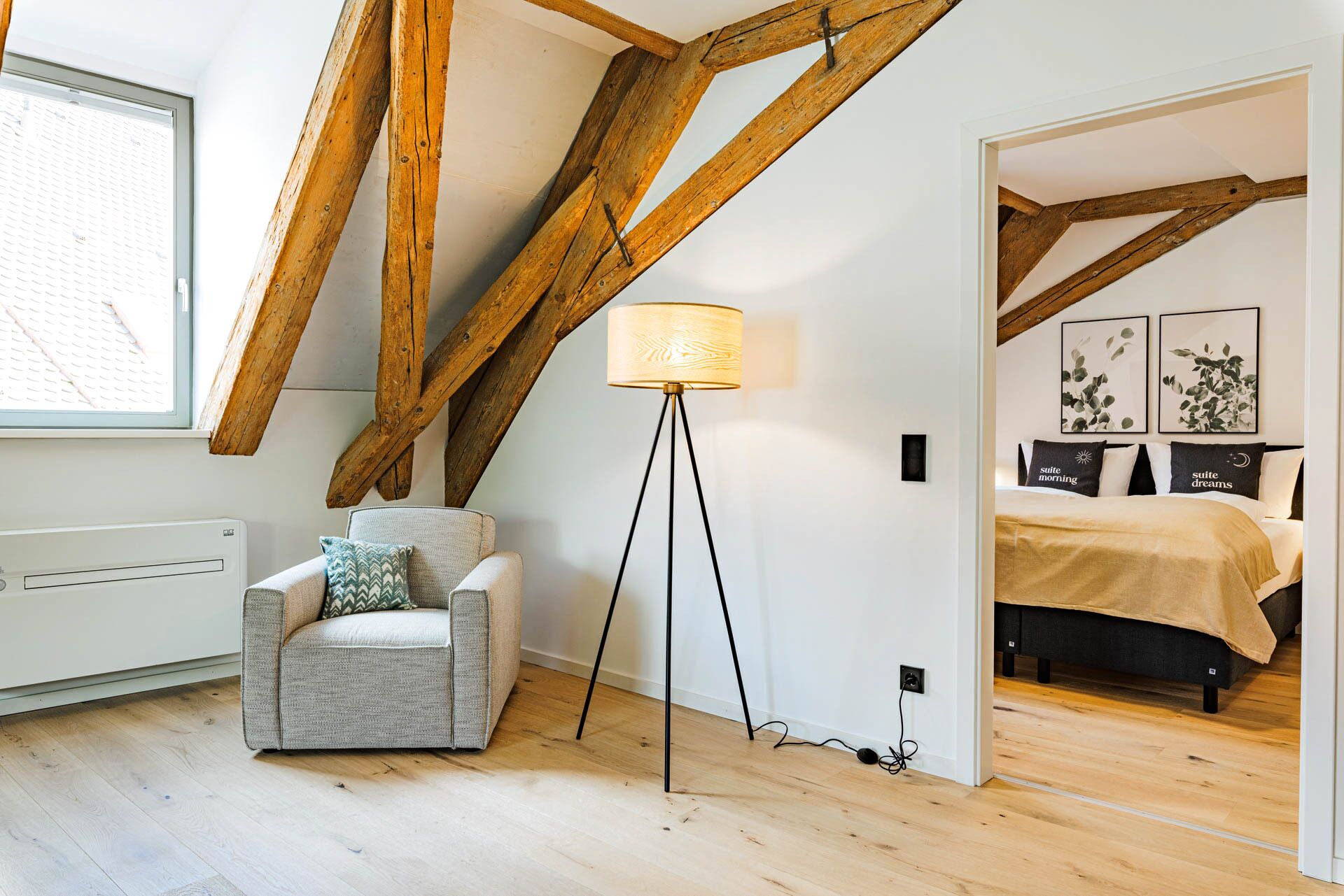 A bedroom with a bed, a chair, a floor lamp, and wooden beams on the ceiling.