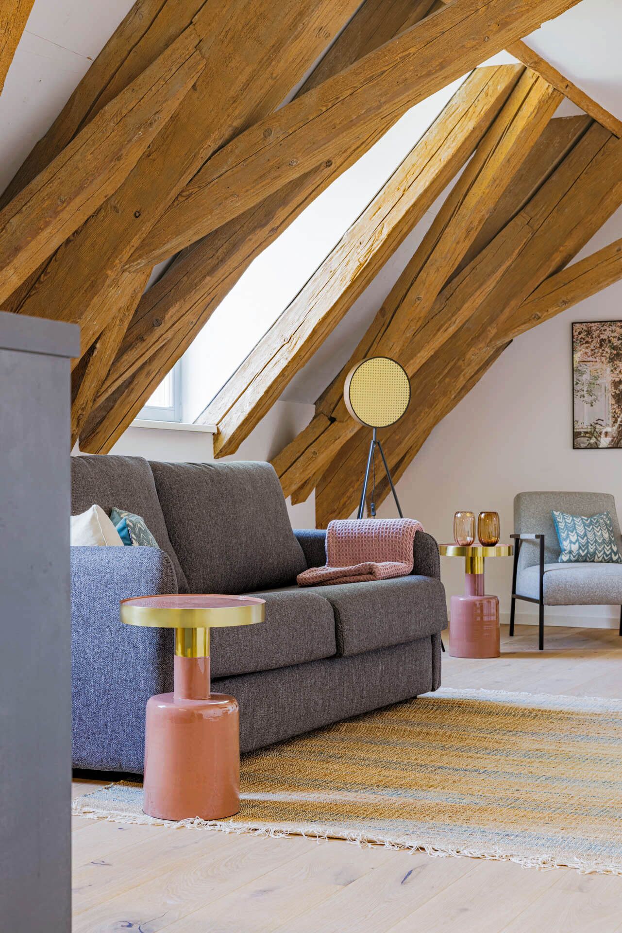 Un espacio tipo loft con vigas de madera a la vista, un sofá gris, una mesa redonda con patas rosas y una alfombra estampada.