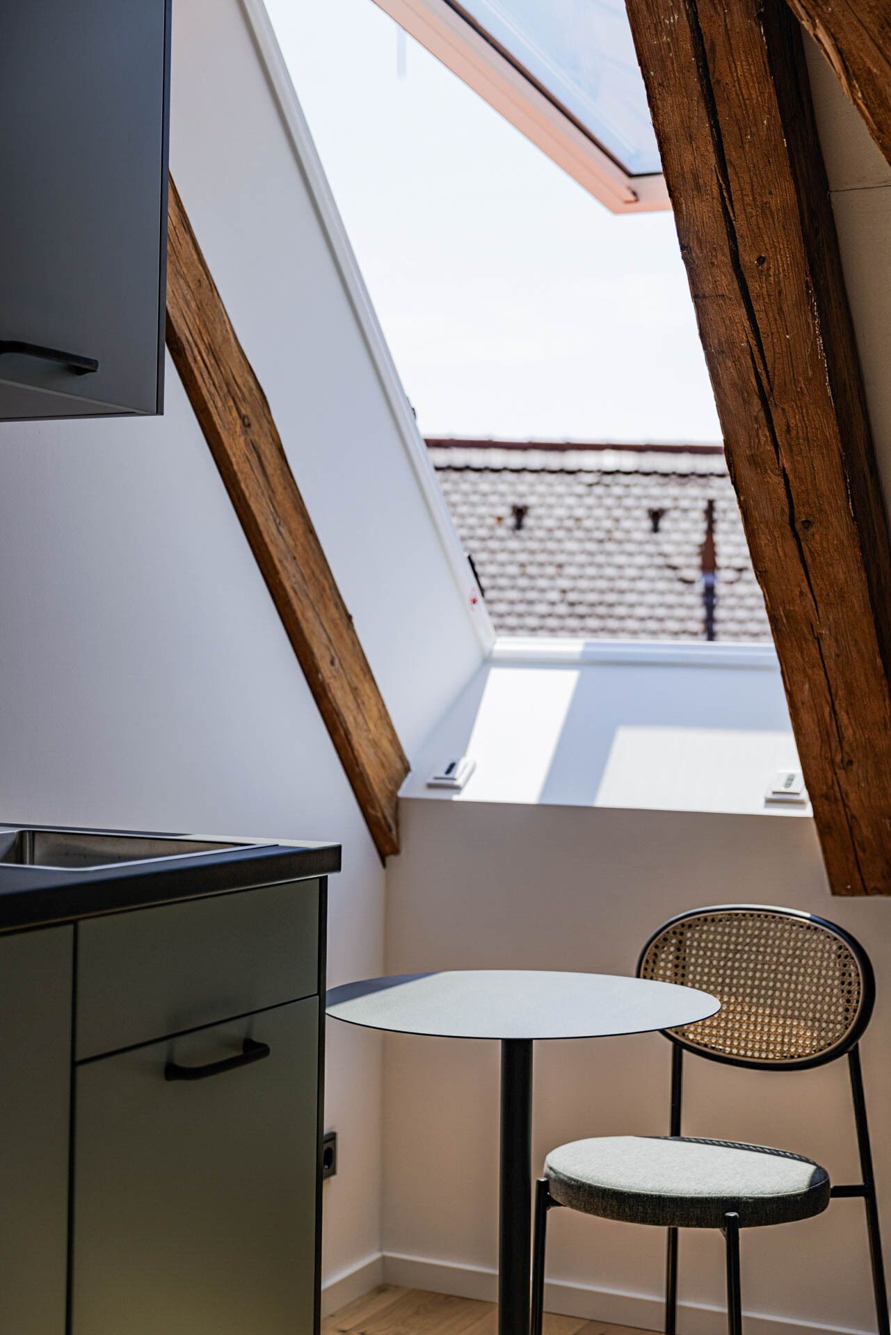 Un petit balcon fermé, équipé d’une table et d’une chaise, avec une poutre en bois et une vue sur un immeuble dont le toit est recouvert de tuiles.