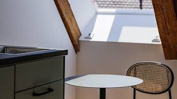 Un petit balcon fermé, équipé d’une table et d’une chaise, avec une poutre en bois et une vue sur un immeuble dont le toit est recouvert de tuiles.