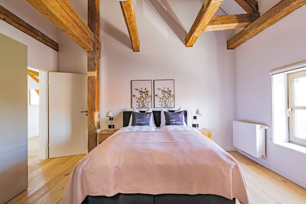 Une chambre avec un grand lit, des poutres en bois, un radiateur et deux œuvres d’art encadrées accrochées au mur.