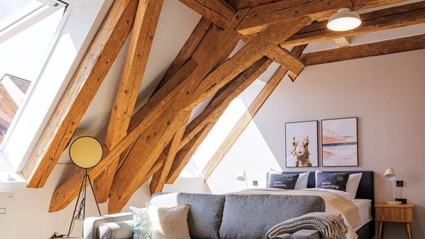 Une chambre à coucher aménagée dans un espace de type « loft », avec des poutres en bois apparentes, un canapé gris, un lit doté d’une tête de lit et une grande lucarne.