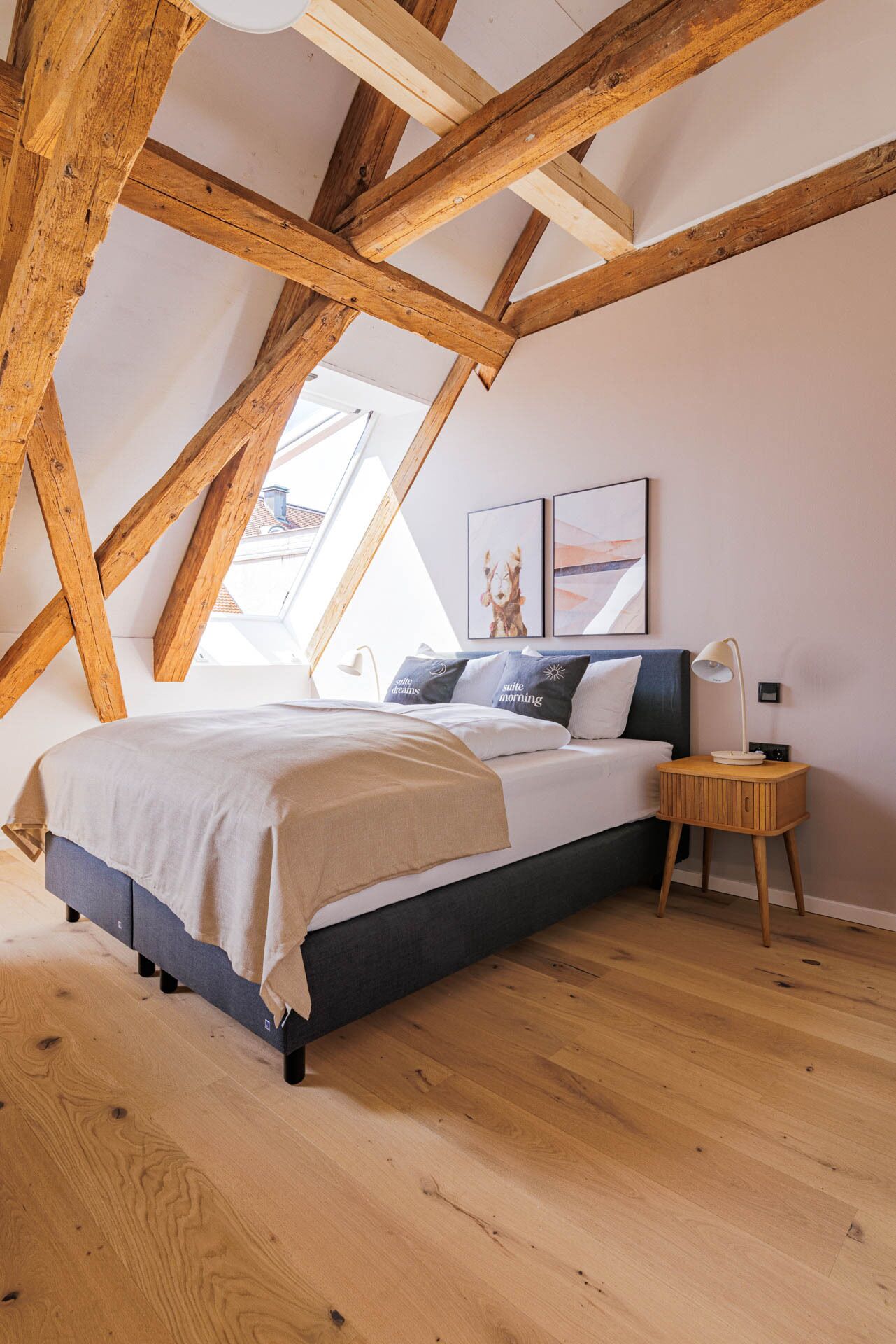 A bedroom with a sloped ceiling, wooden beams, a bed with a grey and white duvet, a bedside table with a lamp, and two framed pictures on the wall.