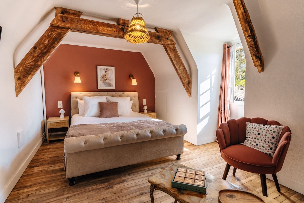 A bedroom with a large bed, wooden beams, a hanging light fixture, and a view of the outdoors.
