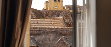 Une vue à travers une fenêtre ouverte, donnant sur un bâtiment doté d’une tour horlogée, d’une porte en bois et d’un cactus.