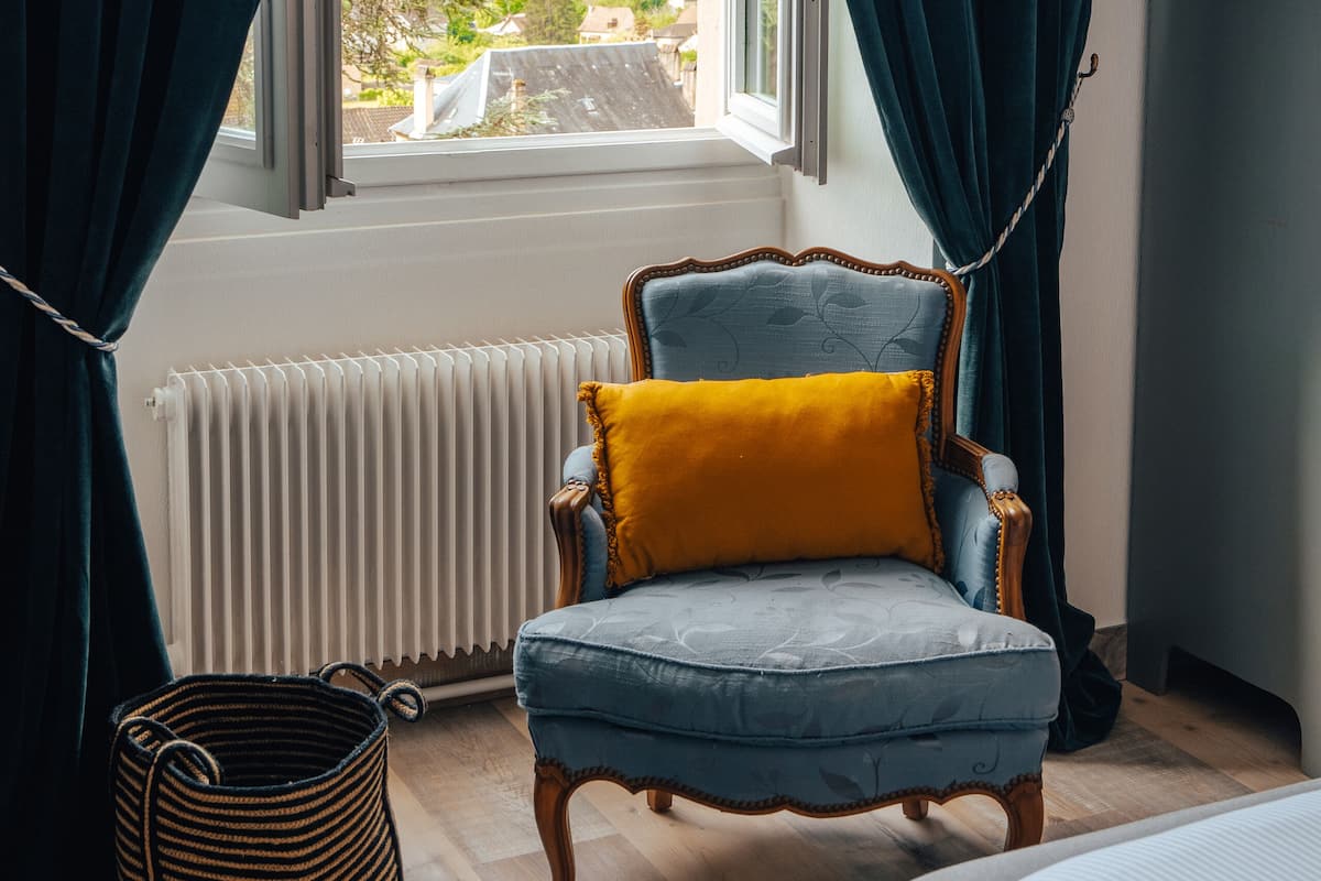 A room with a view of a town, a blue armchair with a yellow pillow, a radiator, and a wooden floor.