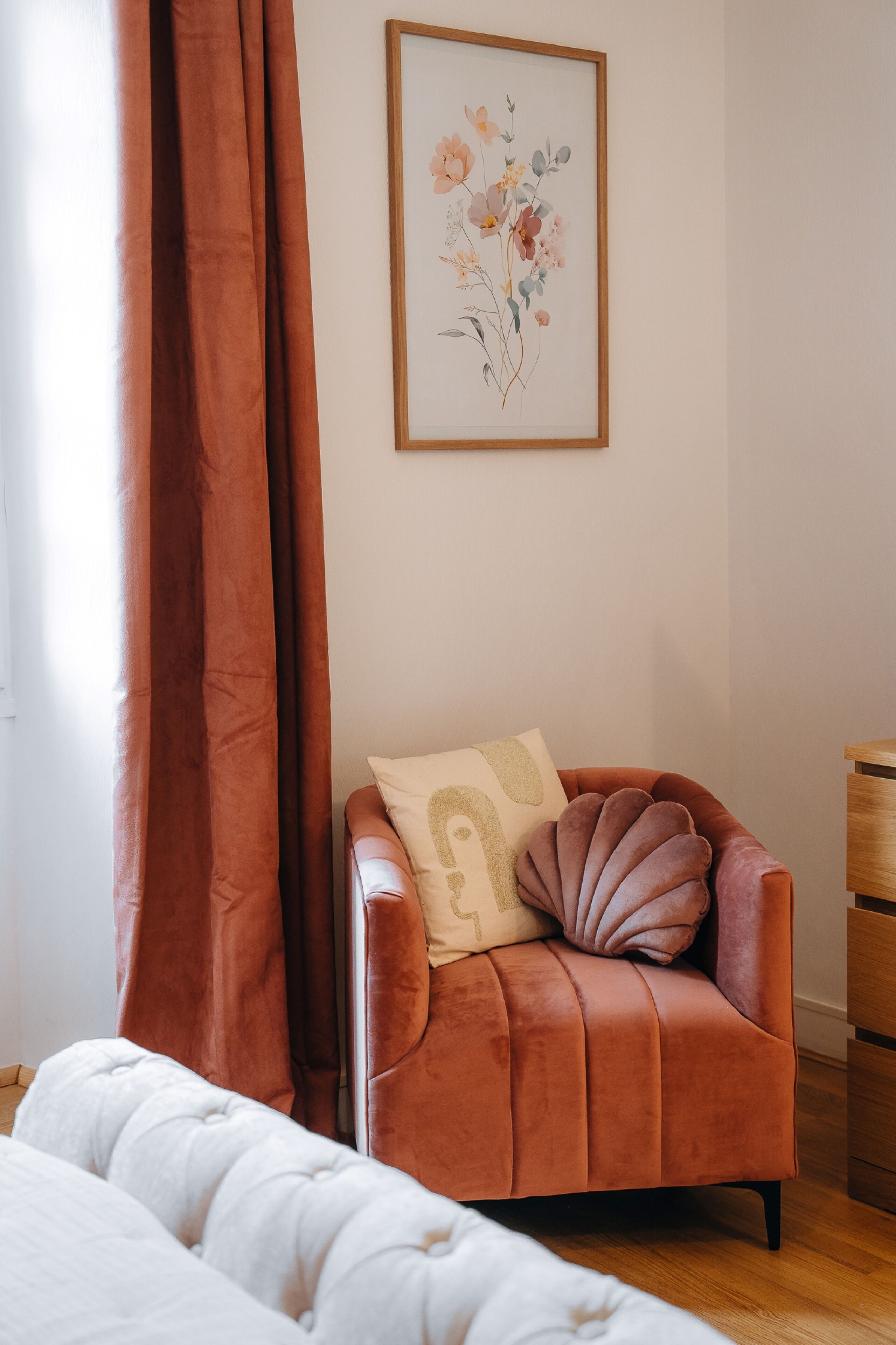 Un dormitorio con cama, un sillón marrón con cojín, una obra de arte floral enmarcada en la pared y una mesita de noche de madera.