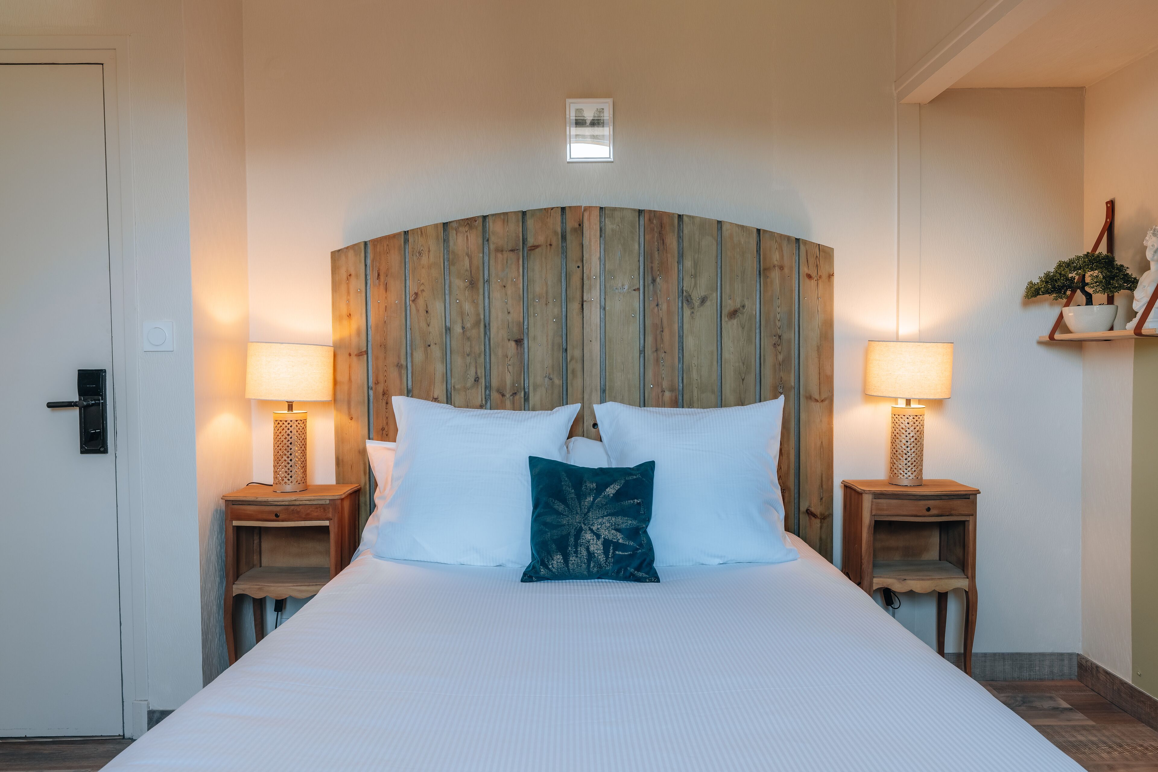 A large bed with white bedding, two bedside tables with lamps, a wooden headboard, and a small shelf with a plant.