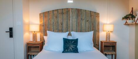 A large bed with white bedding, two bedside tables with lamps, a wooden headboard, and a small shelf with a plant.