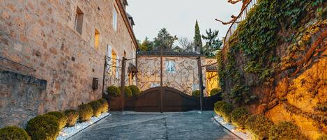 A stone building with a gated entrance, a paved pathway, and a row of bushes along the sides.
