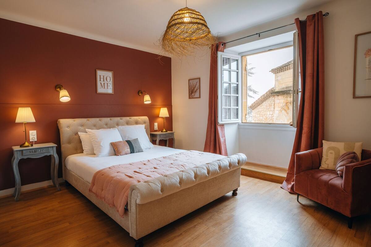 A bedroom with a large bed, two bedside tables with lamps, a window with curtains, and a view of buildings outside.