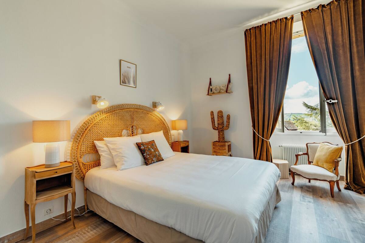 A well-lit bedroom with a large bed, two bedside tables, a chair, and a window with curtains.