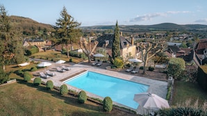 A swimming pool surrounded by lounge chairs and umbrellas, with a view of a village and hills in the background. - Hotel L'abbaye (Saint-Cyprien)
