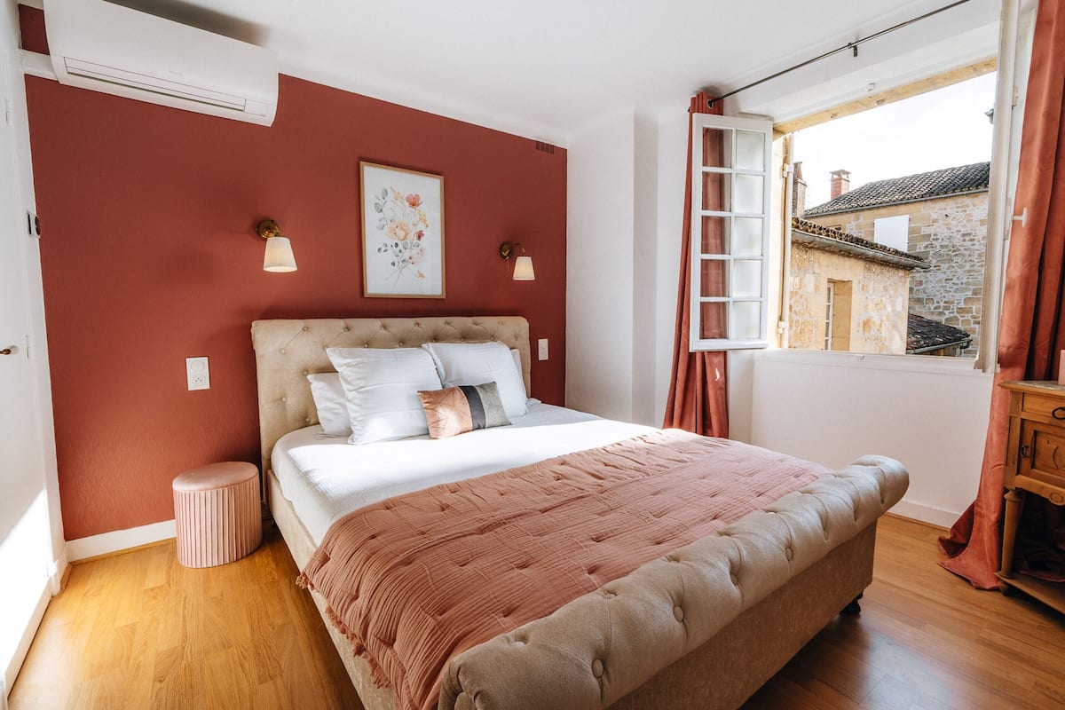 A bedroom with a large bed, a small stool, a window with curtains, and a wall-mounted air conditioner.