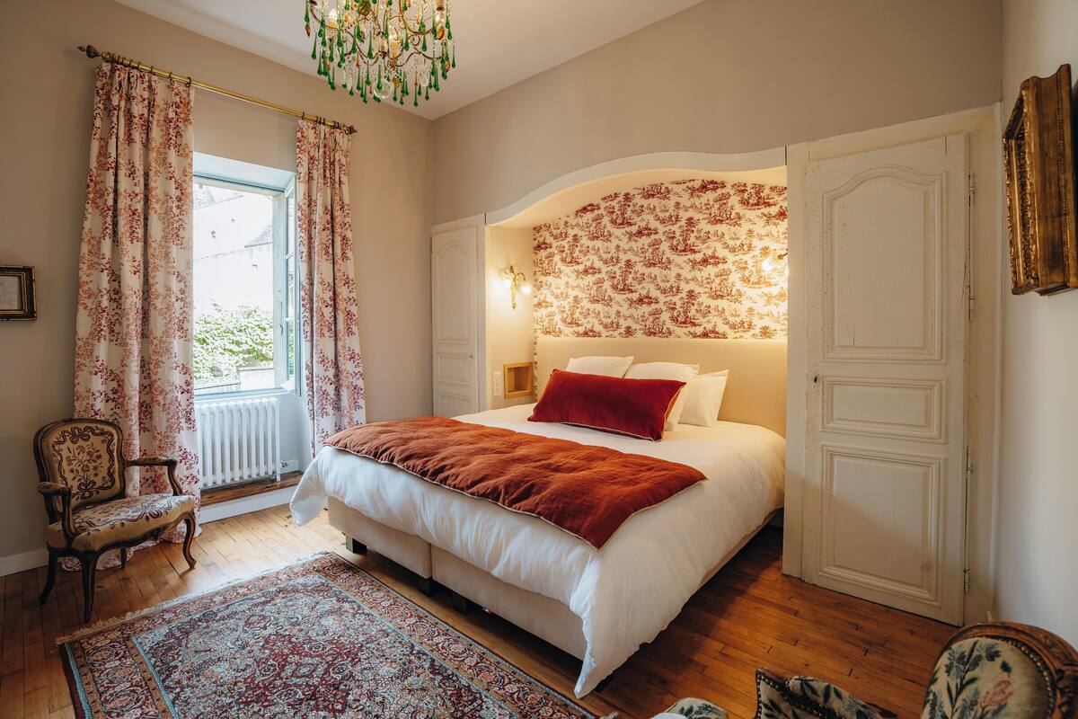 A bedroom with a large bed, a chair, a radiator, and a window with curtains.